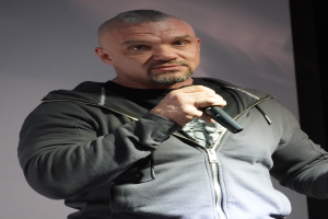 Ein Mann in einer schwarzen Kapuzenjacke und einem schwarzen Anzug mit einem weißen Hemd, glattem Haar und einem selbstbewussten Gesichtsausdruck hält ein Mikrofon, während er vor einer Wand steht.