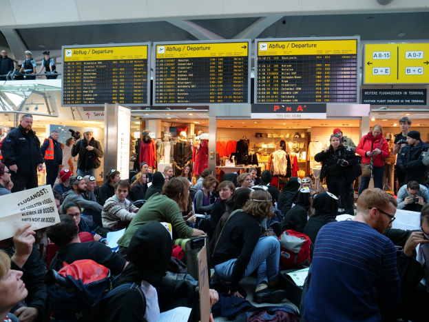 Eine große Gruppe von Menschen sitzt und steht in einem Flughafen während einer Protestaktion, mit Informationsschildern, Schaufensterpuppen in Kleidern und Deckenbeleuchtung im Hintergrund.