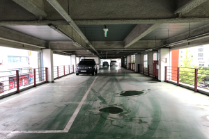 Ein mehrstöckiges Parkhaus voller geparkter Autos, umgeben von Geländern, Säulen, Deckenleuchten und sichtbaren Rohren, mit Bäumen und einer Stadtkulisse von Gebäuden mit Fenstern im Hintergrund.