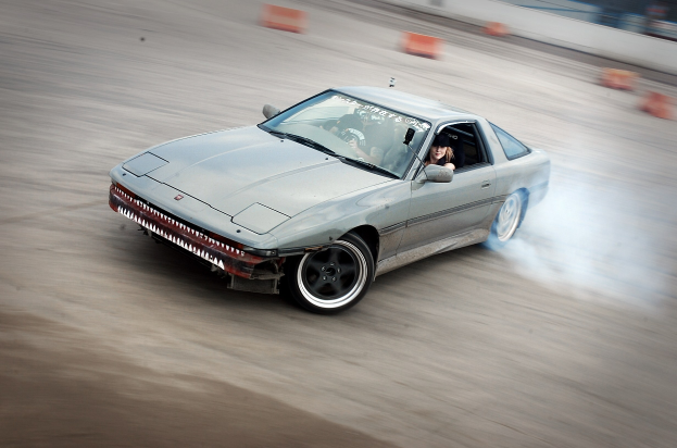 Eine Person mit Helm fährt ein Auto auf einer Strecke, aus dem Rauch austritt, umgeben von orangen Konen mit einem unscharfen Hintergrund, der hohe Geschwindigkeit andeutet.