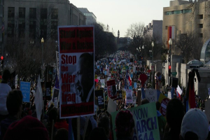 Eine Gruppe von Menschen demonstriert auf einer Straße, hält Plakate, mit Gebäuden, Bäumen, Laternenmasten und dem Himmel im Hintergrund.