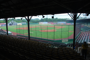 Ein Stadion mit Stühlen im Vordergrund, ein Baseballfeld in der Mitte, Bäume, Banner und ein Scoreboard im Hintergrund, unter einem bewölkten Himmel.