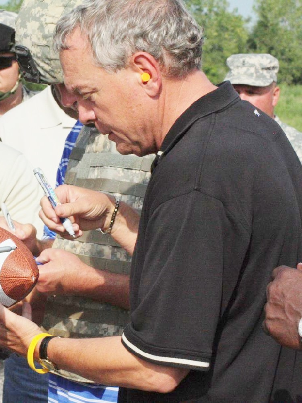 Ein Mann in schwarzem Hemd unterschreibt einen Fußball, während er von einer Gruppe von Menschen umgeben ist, die Kappen und Schutzbrillen tragen. Im Hintergrund sind Bäume und ein klarer blauer Himmel zu sehen.