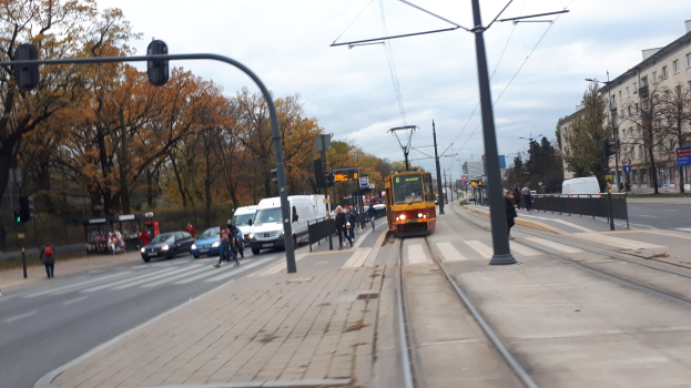 Eine Stadtstraße mit einer gelben Straßenbahn auf den Gleisen, Fahrzeugen, Fußgängern, Strommasten mit Leitungen, Verkehrsampeln, Hinweisschildern, Bäumen, Gebäuden mit Fenstern und einem bewölkten Himmel.