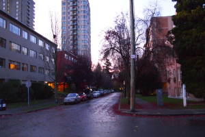 Eine Straßenszene vor einer Stadt mit Gebäuden, Bäumen und Autos unter einem bewölkten Himmel.