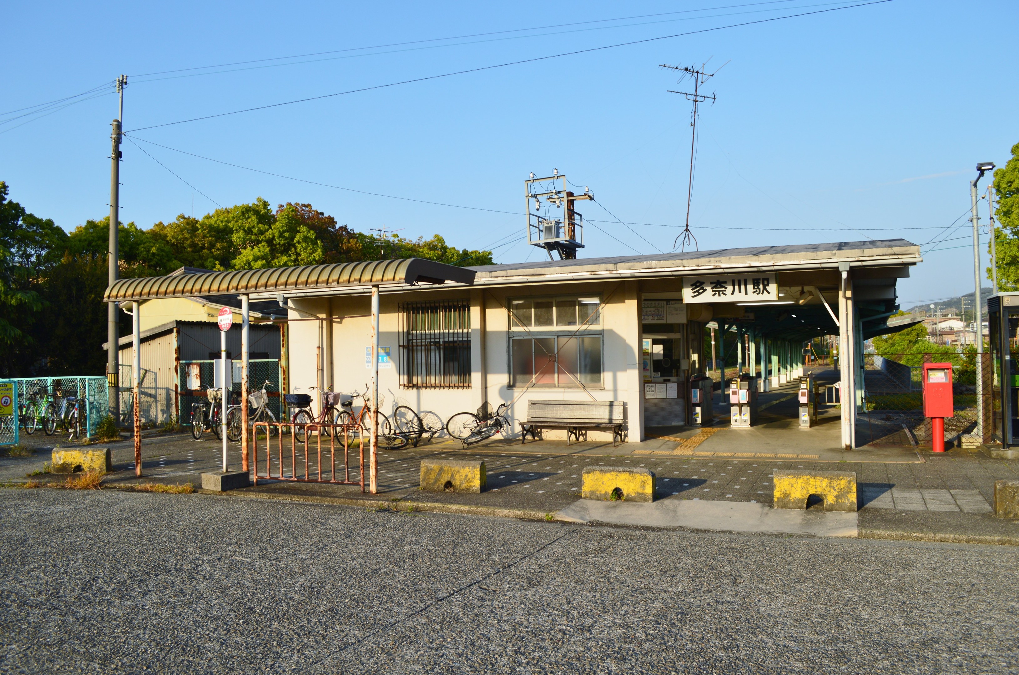 Ein Bahnhof mit vor ihm geparkten Fahrrädern, eine Straße unten und Häuser, Bäume, Pfähle, Drähte und einen klaren blauen Himmel im Hintergrund.