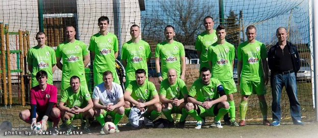 Eine Gruppe von Männern in grünen T-Shirts steht auf einem Fußballfeld in der Nähe eines Torpfostens, einige hocken, mit einem Ball davor, Bäume und Gebäude im Hintergrund.