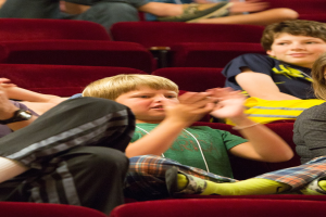 Eine Gruppe von Menschen in einem Kino, die applaudieren, mit einem Jungen in der Mitte, umgeben von sitzenden Personen.