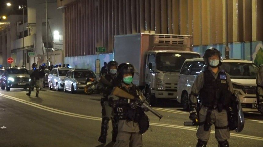 Eine Gruppe von Polizisten in Helmen, Handschuhen und Gesichtsmasken steht an einer Straße in der Nacht, einige halten Waffen, mit Fahrzeugen, Laternenmasten, Schildern und Gebäuden im Hintergrund.