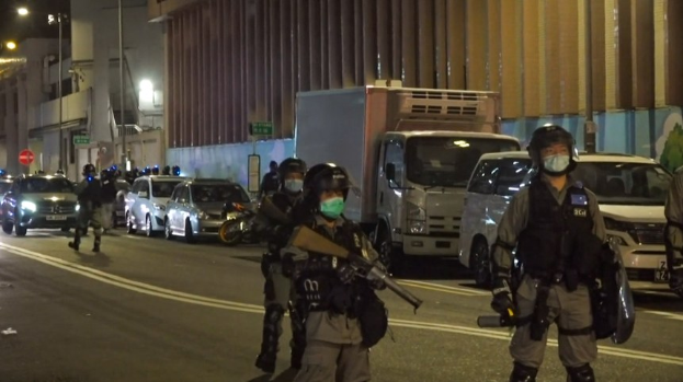 Eine Gruppe von Polizisten in Helmen, Handschuhen und Gesichtsmasken steht an einer Straße in der Nacht, einige halten Waffen, mit Fahrzeugen, Laternenmasten, Schildern und Gebäuden im Hintergrund.