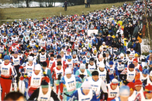 Eine große Gruppe von Menschen, die bei einem Rennen auf Schnee laufen, tragen Mützen und halten Ski-Stöcke, mit Bäumen im Hintergrund und Text unten.