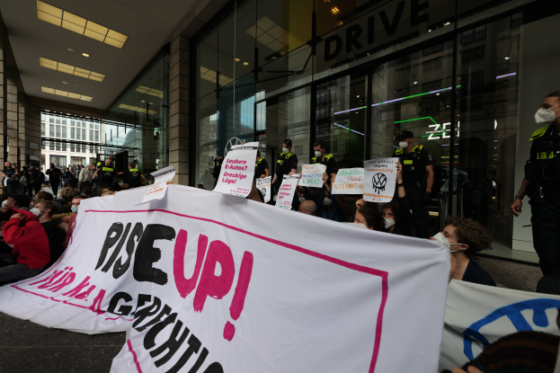 Eine Gruppe von Menschen hält ein Transparent und Schilder mit dem Text "Erhebt euch" und protestiert vor einem Gebäude mit Glaswänden und Deckenleuchten, einige tragen Mäskchen.