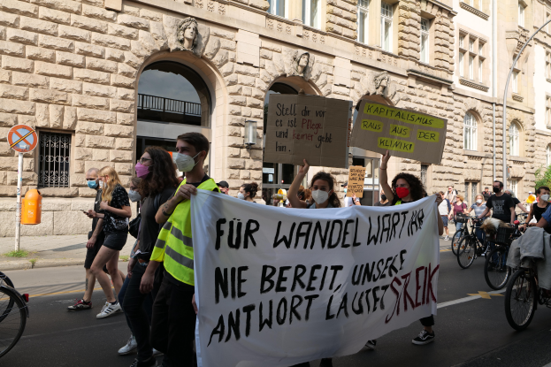 Eine Gruppe von Menschen marschiert auf einer Straße in Berlin, wobei einige Plakate und Banner halten und andere Fahrräder fahren, vor einem Gebäude mit Bögen, Säulen, Skulpturen und Bäumen.