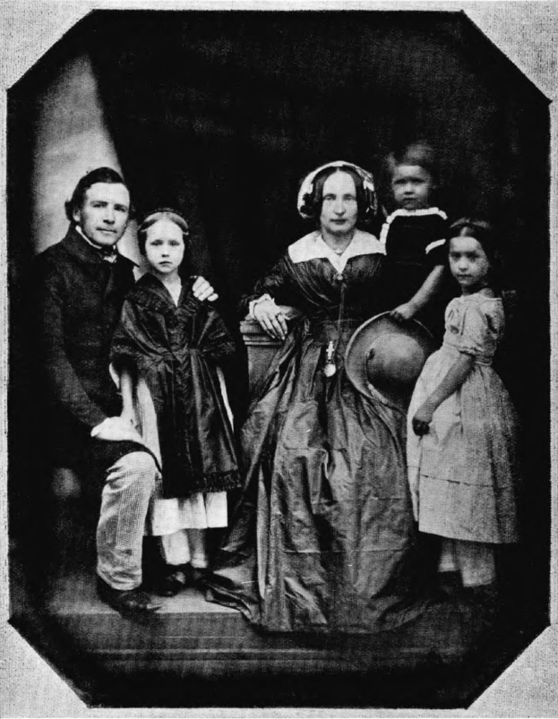 Ein Schwarz-Weiß-Foto einer Familie, die posiert, mit einer Frau in einem Kleid, die einen Hut hält, zwei Kindern neben ihr und einer Treppe, die zu ihnen führt.