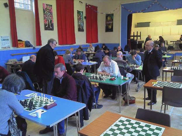 Ein großer Saal mit zwei Personen, die an einem Tisch Schach spielen, mit Münzen auf dem Schachbrett, Vorhängen, Plakaten, einem Fenster und einem Kalender an der Wand.