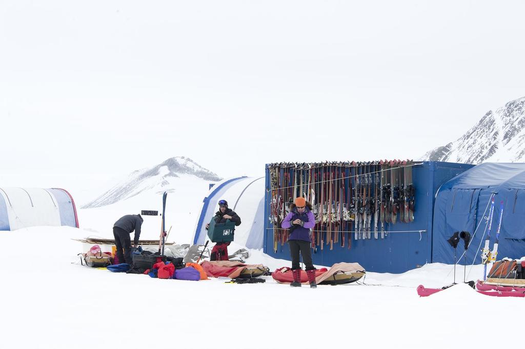Drei Personen auf einer schneebedeckten Landschaft mit verstreuten Taschen, Zelten mit Skiern darauf und schneebedeckten Hügeln im Hintergrund unter einem klaren Himmel.