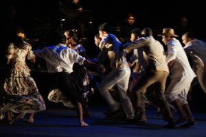 Eine Gruppe von Menschen tanzt im Vordergrund mit einer anderen Gruppe von Menschen, Ständen und Mikrofonen im Hintergrund, alles auf einem Boden.