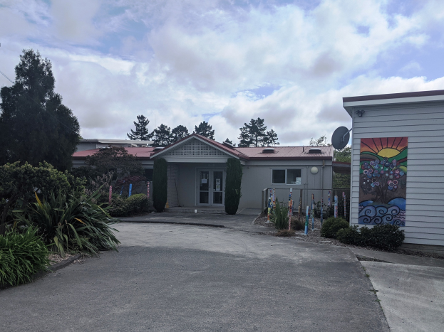 Eingang eines Kinderspielplatzes mit einer Wandmalerei an der Seite, umgeben von Grün, einer Straße im Vordergrund und einem bewölkten Himmel darüber.