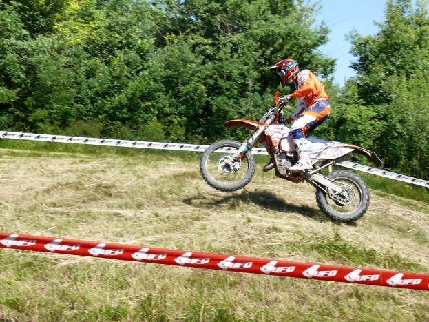 Ein Radfahrer mit Helm fährt an Absperrband vorbei, im Hintergrund sind Bäume, Gras und Himmel zu sehen.