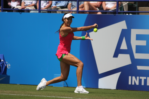 Eine Frau spielt Tennis, hält einen Schläger in ihrer rechten Hand, trägt eine weiße Mütze und Schuhe, hat ein grünes Band am linken Handgelenk und befindet sich auf einem grünen Platz mit Zuschauern im Hintergrund.