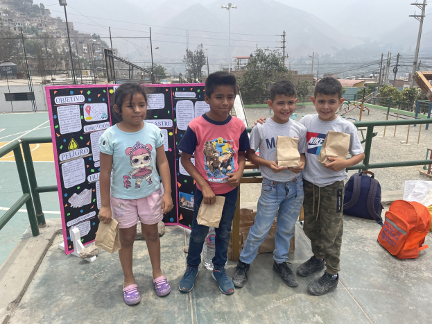 Gruppe von Kindern auf einem Basketballfeld, die Papiertüten halten, mit Gegenständen auf der rechten Seite und einer Tafel mit Text auf der linken Seite, vor dem Hintergrund von Zaun, Pfosten, Straßenlaternen, Bäumen, Gebäuden, Hügeln und Himmel.