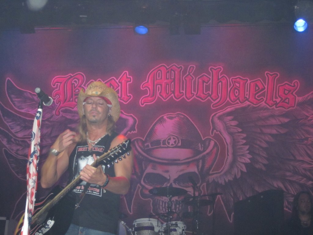 Ein musikalisches Konzert-Szenario mit einem Gitarristen links neben einem Mikrofon, einer Person, die unten rechts sitzt, einem Banner im Hintergrund und Drogen in der unteren Mitte, alles unter Bühnenbeleuchtung.