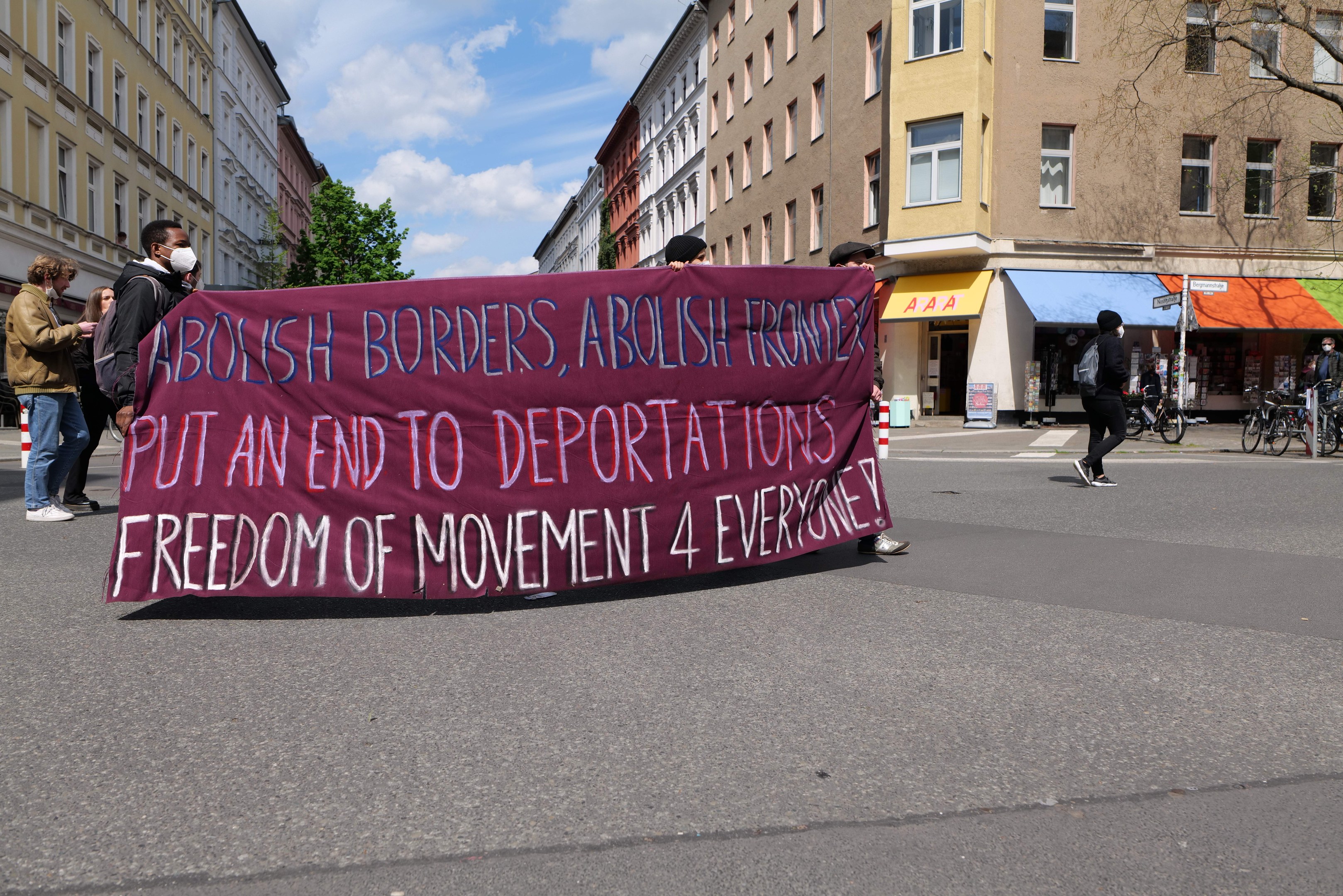 Eine Gruppe von Menschen marschiert mit einem Banner, auf dem "Abolish Borders, Abolish Frontiers, Put an End to Deportations, Freedom of Movement 4 Everyone" steht, durch eine Straße, mit Gebäuden, Bäumen, Fahrrädern und einem bewölkten Himmel im Hintergrund.