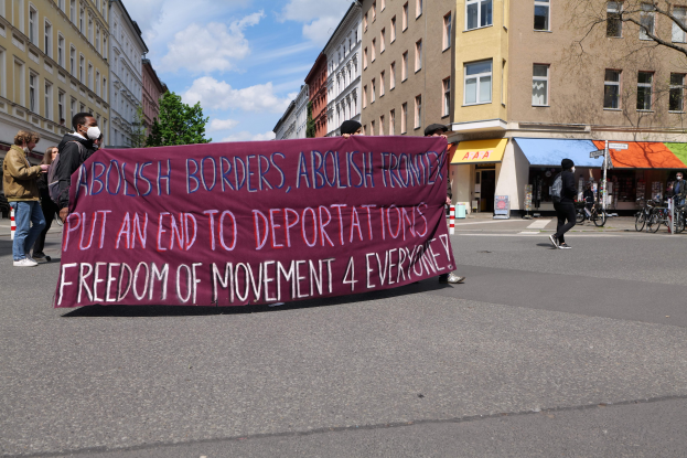 Eine Gruppe von Menschen marschiert mit einem Banner, auf dem "Abolish Borders, Abolish Frontiers, Put an End to Deportations, Freedom of Movement 4 Everyone" steht, durch eine Straße, mit Gebäuden, Bäumen, Fahrrädern und einem bewölkten Himmel im Hintergrund.