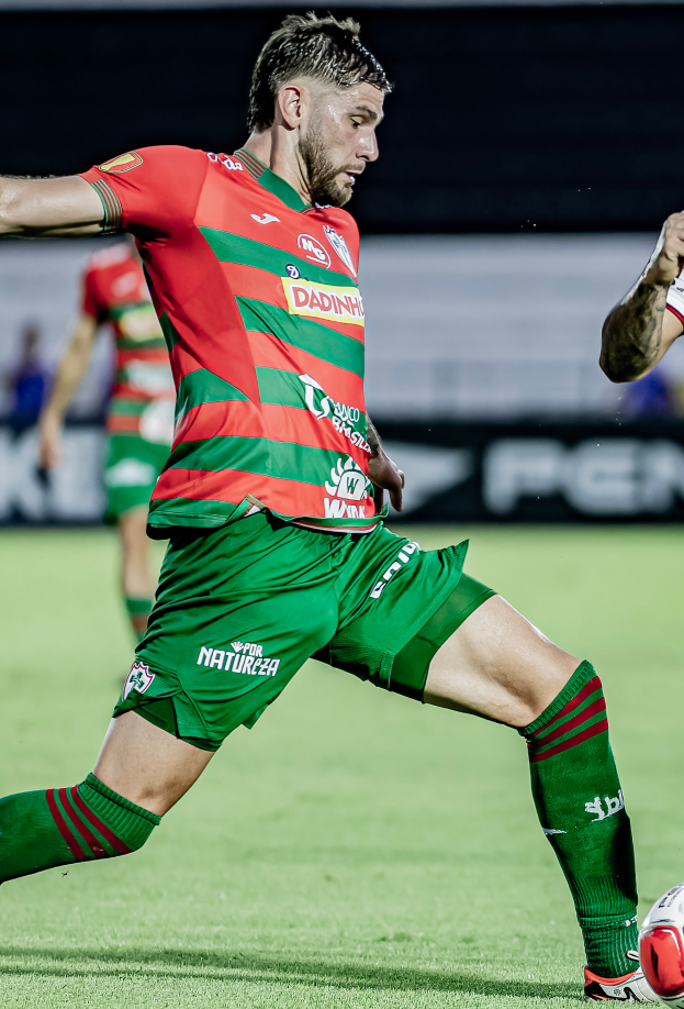Ein Mann in roter und grüner Uniform tritt einen Fussball auf einem Rasenfeld, mit einer anderen Person im Hintergrund und einer Tafel, auf der "Santos vs Vitoria Guimaraes" steht.