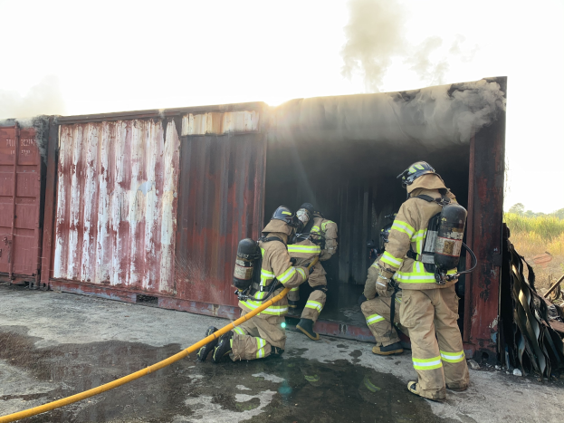 Feuerwehrleute in Schutzausrüstung löschen ein Feuer in einem Seecontainer, mit Rauchentwicklung und Grünfläche im Hintergrund.