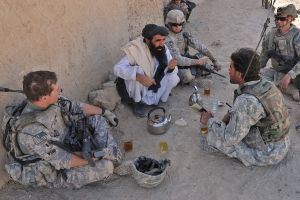 Eine Gruppe von Menschen, einer in einem weißen Hemd und andere in Armeeuniformen mit Waffen, die um einen Tisch mit einem Kessel und Gläsern sitzen, mit einer Wand auf der linken Seite.