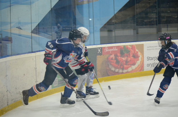 Gruppe junger Menschen, die Eis hockey auf einer Indoor-Eisfläche spielen, mit Helmen, Sportuniformen und Schlittschuhen sowie Hockeystöcken in der Hand, vor einer Glaswand und einem Plakat im Hintergrund.