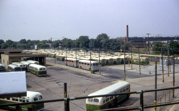 Eine Reihe geparkter Busse auf einer Straße mit Polen, Drähten, Gebäuden, Bäumen, einem Zaun und einem Rauchschlot unter dem Himmel.