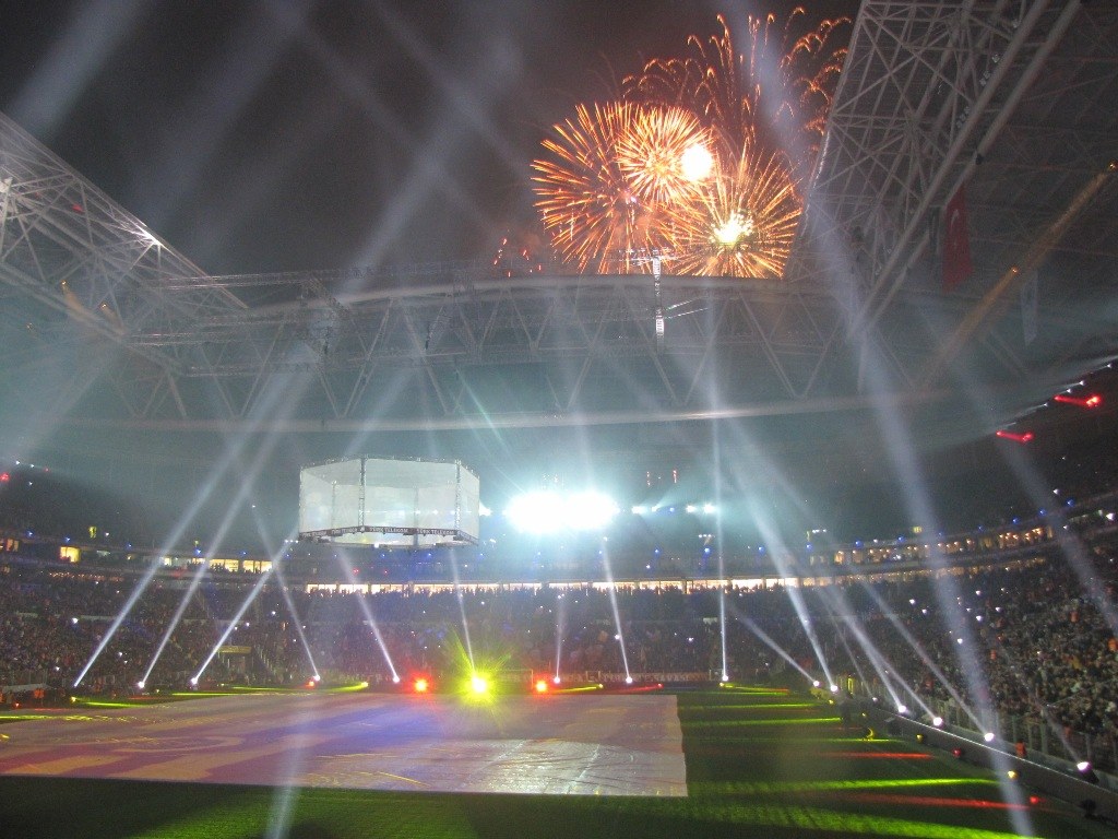Das Olympische Stadion in London, England, erleuchtet nachts mit hellen Lichtern, umgeben von einer großen Menge, grünem Gras und Feuerwerk, das den Himmel erhellt.