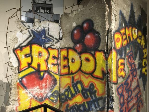 Mauer mit Graffiti, das in fetten schwarzen Buchstaben "Freiheit und Demokratie" liest, einem Geländer unten und einem Gebäude mit Fenstern im Hintergrund.