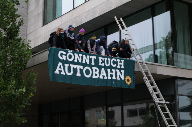 Gruppe von Menschen auf einem Gebäude, das eine Fahne hält, mit einem Baum links und Glasfenstern im Hintergrund, die scheinbar für die deutsche Regierung protestieren.