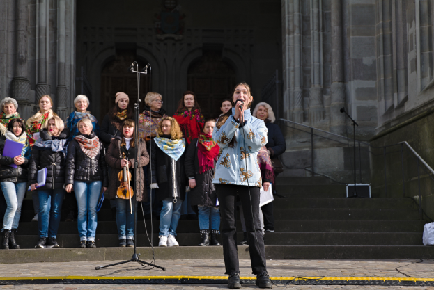 Eine Gruppe von Menschen, einige mit Musikinstrumenten, steht vor einem Gebäude mit Treppe und Geländer, während eine Frau in der Mitte in ein Mikrofon singt, das von Mikrofonstativen umgeben ist.