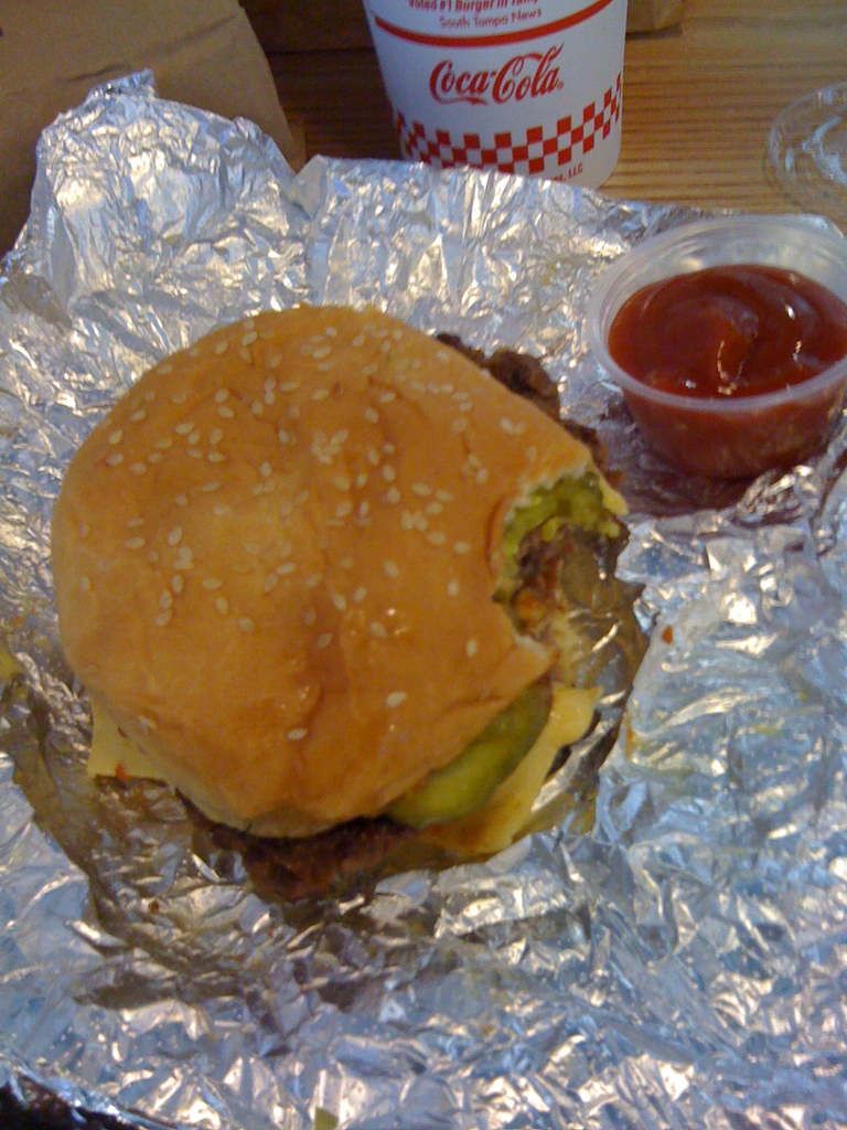 Ein Burger auf Alufolie mit einem Soßenbehälter, einem Becher und anderen Gegenständen auf einem Tisch.