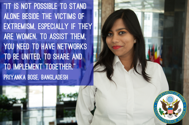 Eine Frau in einem weißen Hemd steht vor einem Gebäude mit einer Aussage über Einheit und Unterstützung von Opfern von Extremismus, insbesondere Frauen, mit einem Logo in der unteren rechten Ecke.