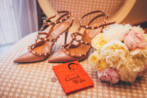 Ein Tisch zeigt ein Buch mit dem Titel "Wie werde ich eine gute Ehefrau", einen Blumenstrauß und ein Paar Valentino-Schuhe vor einem Vorhang.