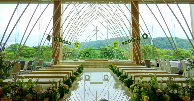 Eine Hochzeitszeremonie in einem großen Saal mit halbrunder Bestuhlung, geschmückt mit Blumenbouquets und anderen Gegenständen, aus Bodensicht mit Bäumen, Hügeln und einem klaren blauen Himmel im Hintergrund.