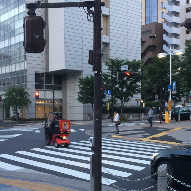 Ein Roller ist auf der Straße mit ein paar Menschen im Hintergrund, eine Ampel im Vordergrund und Gebäude mit Bäumen in der Ferne.