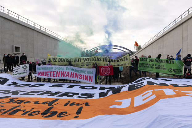 Eine Gruppe von Menschen steht vor einer großen Plane mit der Aufschrift "Klima Protest in Berlin", hält Schilder und Fahnen und hat Rauch im Hintergrund und einen bewölkten Himmel über sich.