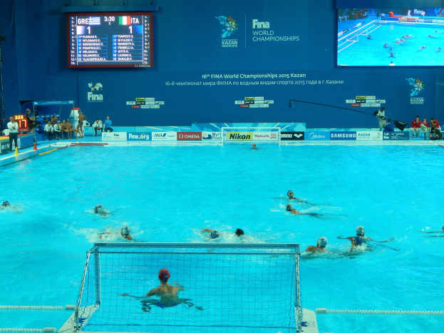 Eine Gruppe von Menschen spielt Wasserball in einem Schwimmbecken während der FINA-Weltmeisterschaften 2016 in Kazan, mit Zuschauern auf der linken Seite und einem Bildschirm im Hintergrund, der das Event-Branding anzeigt.