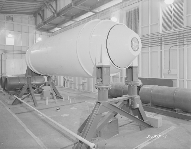 Schwarzes und weißes Foto einer großen zylindrischen Wasserstoffbombe auf einem Ständer in einer Fabrik mit Rohren, Lampen und einer Wand im Hintergrund und Text unten.