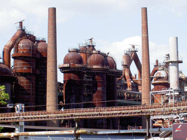 Großes industrielles Fabrikgelände mit ausgedehnten Rohrleitungen, mehreren Schornsteinen, einer Brücke, Strommasten mit Laternen, umgeben von Bäumen und einem bewölkten Himmel.