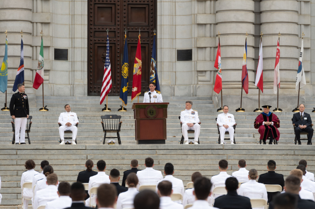 Gruppe von Menschen in weißen Uniformen sitzt auf den Stufen eines Gebäudes mit einem Podium, das von Flaggen flankiert ist, vor einem großen Gebäude mit einer Tür während einer Ehrenzeremonie.