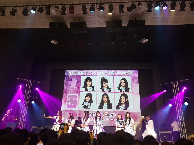 Eine Gruppe von Menschen performs auf einer hell erleuchteten Bühne mit Trägern, als ein Sänger ein Mikrofon hält und ein Musiker ein Instrument spielt, während eine Menge darunter das Ereignis aufzeichnet und ein großer Bildschirm im Hintergrund Bilder der K-Pop-Gruppe BK48 anzeigt.