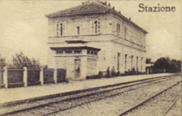 Altes Schwarz-Weiß-Foto einer Bahnstation mit einem zentralen Gebäude, Bäumen, einem Zaun auf der linken Seite, Eisenbahnschienen unten und Text oben.