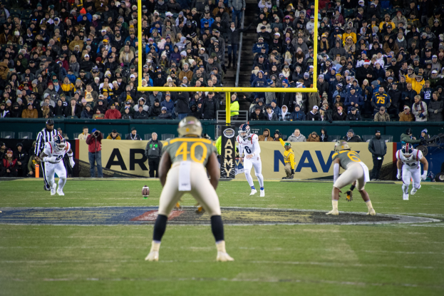 Fußballspieler in Helmen und Uniformen konkurrieren auf einem Feld mit Zuschauern in den Rängen, während Stadionwerbung und Pfosten sichtbar sind.